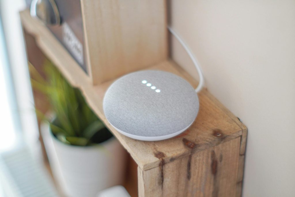 Google Home Mini smart speaker with white LED lights illuminated, sitting on a rustic wooden shelf next to a white planter with green succulent plant