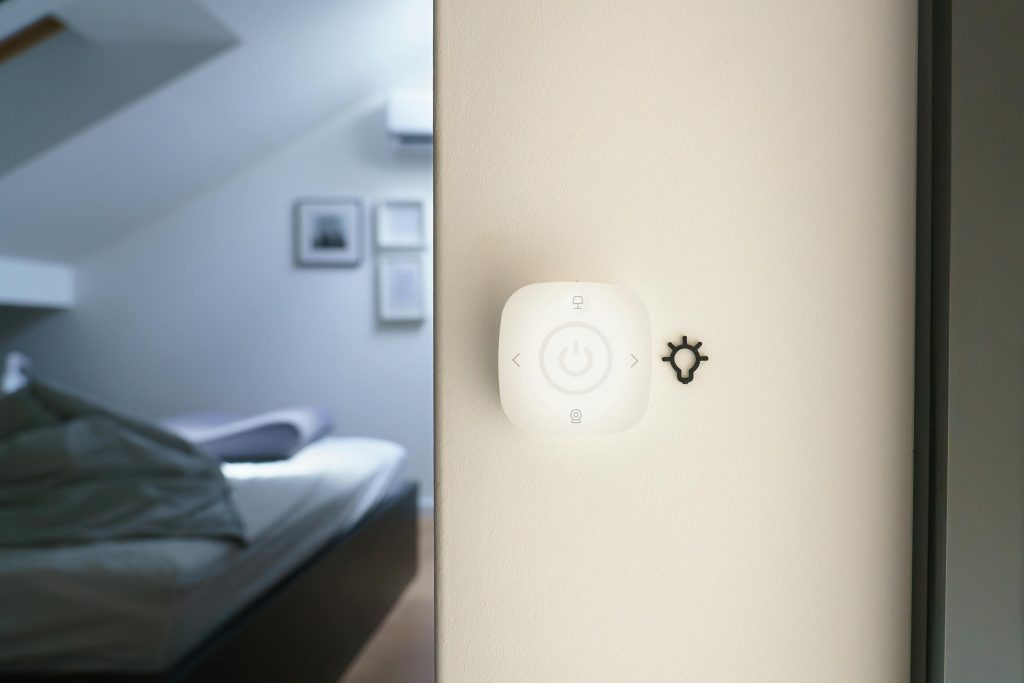 Smart home light control switch mounted on a wall inside a modern, softly lit bedroom.